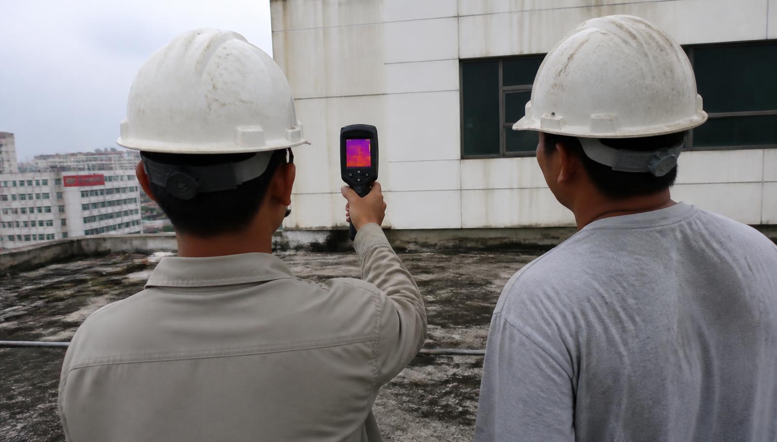 Dois técnicos em cobertura de edifício utilizando câmera termográfica na inspeção de fachada